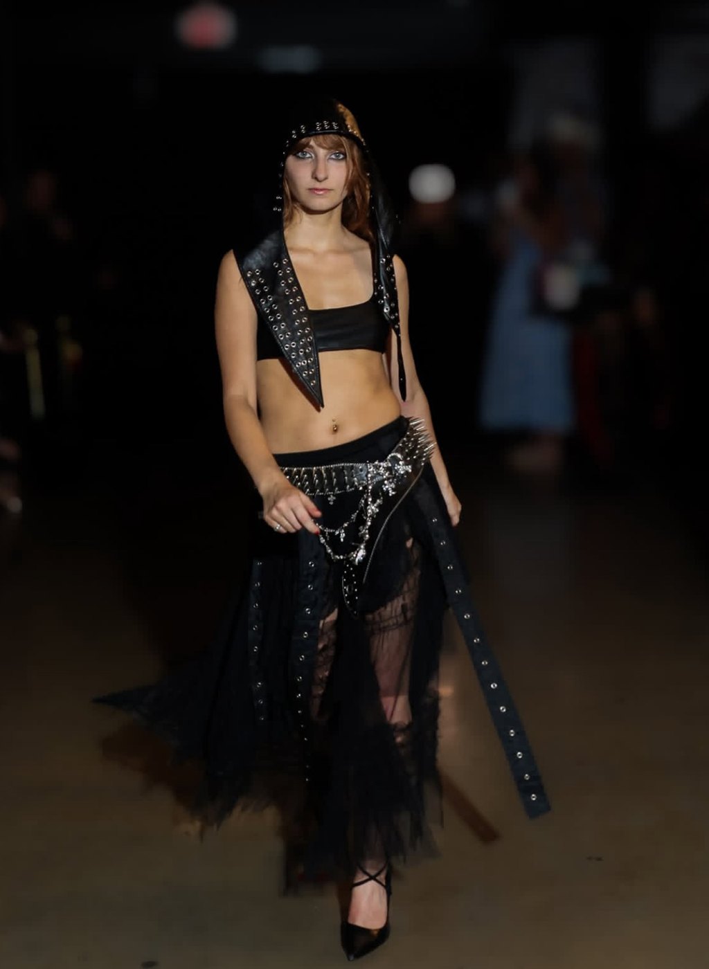 Isabella walking in a studded leather hood, bandeau and tulle skirt with a spiked belt