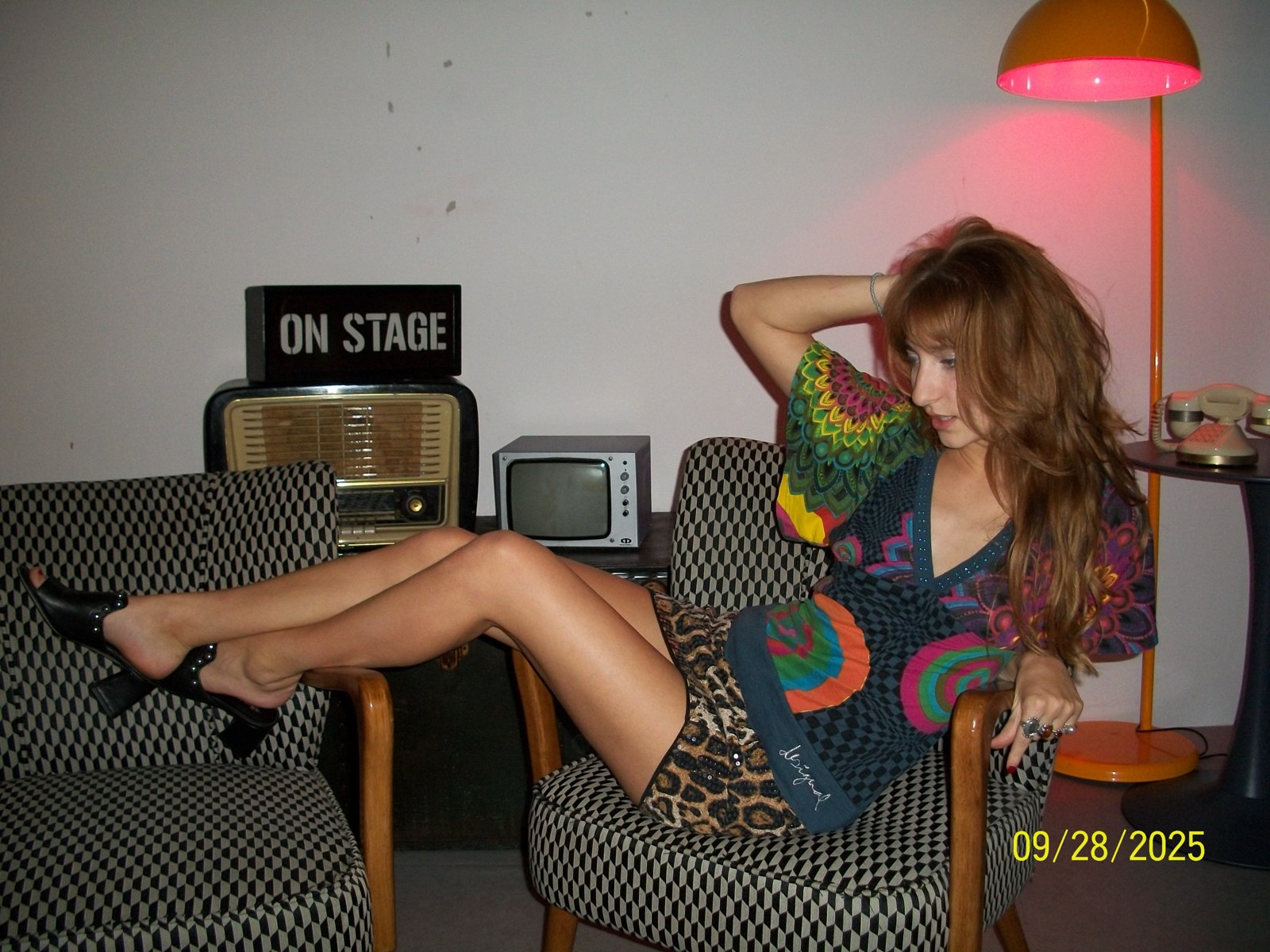 Isabella looking down beside an orange floor lamp with legs crossed over the armchair