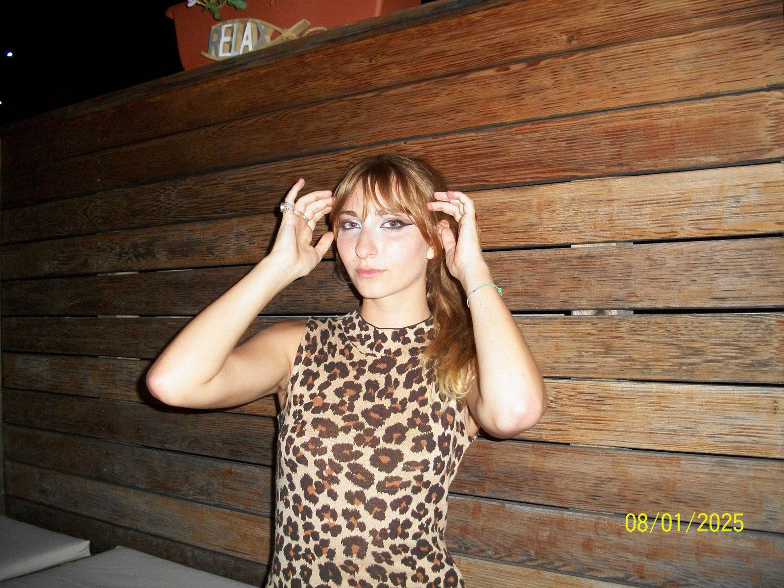 Isabella in a leopard-print sleeveless dress with graphic eyeliner against a wooden wall