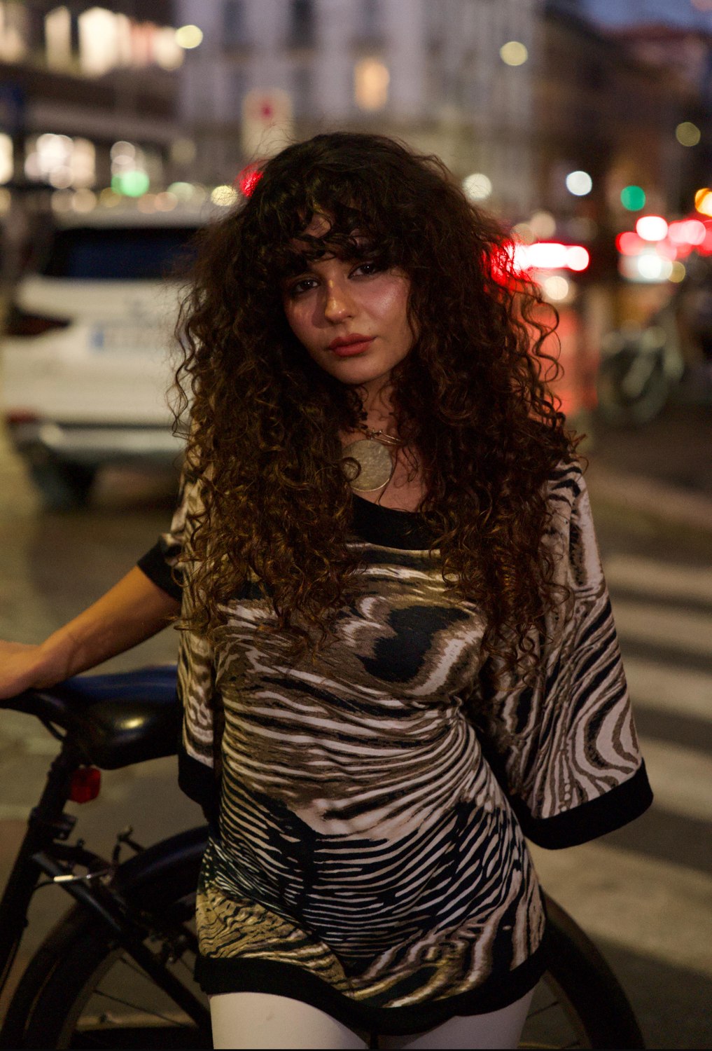 Woman with long curls in an oversized zebra-print tunic and gold pendant on a city street at night