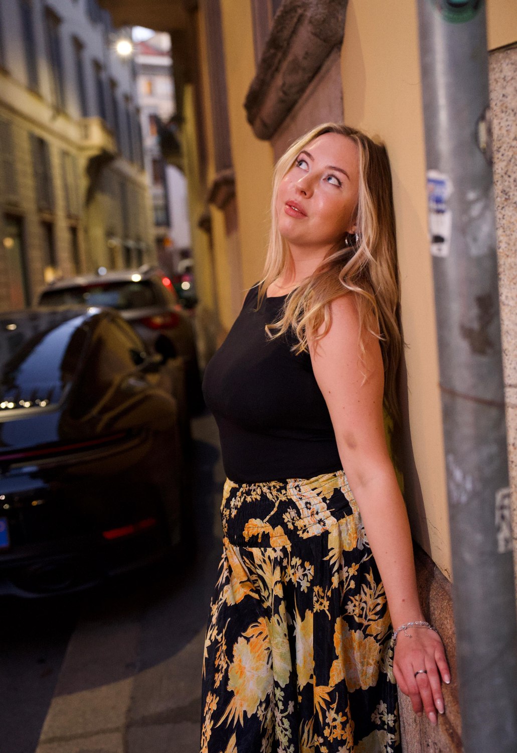 Woman in a black tank and black and yellow floral skirt leaning against a building at dusk