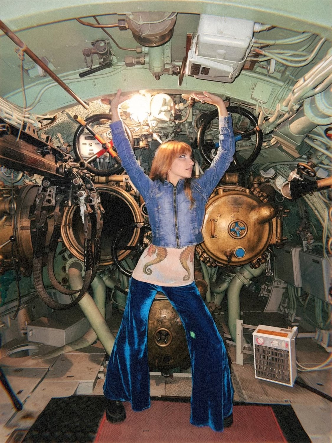Isabella with arms raised in the torpedo room, in a cropped denim jacket, seahorse top and blue velvet flares