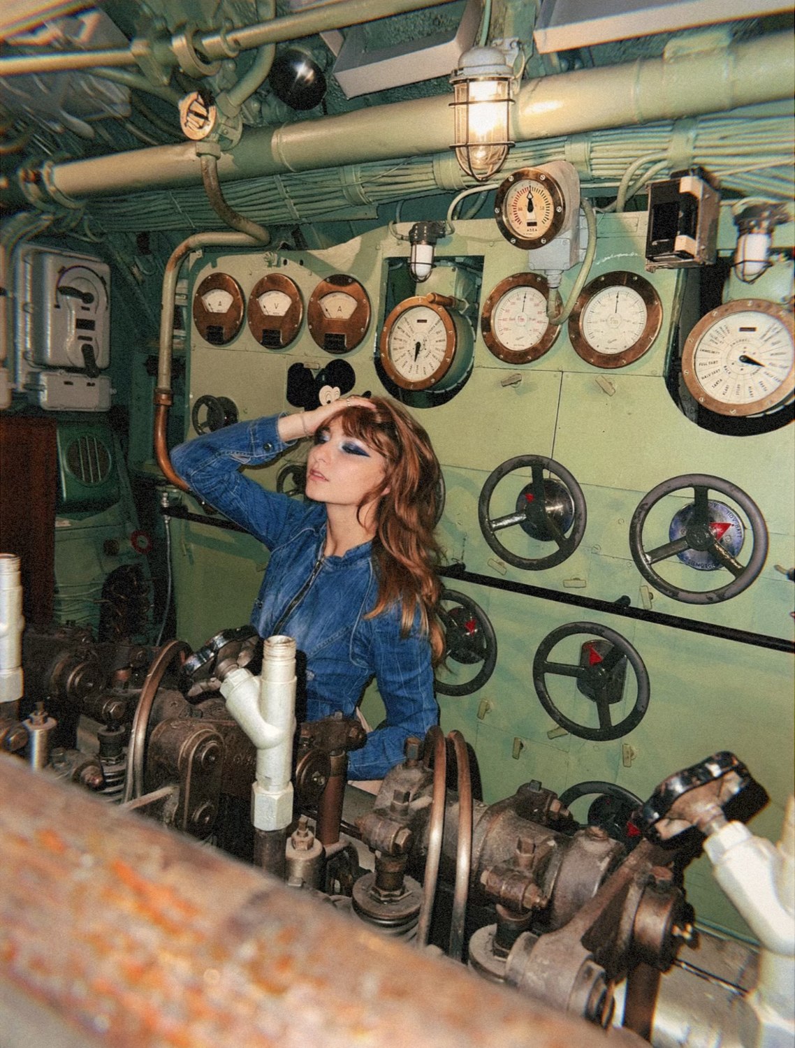 Isabella with a hand in her hair and eyes closed beside the submarine's gauges and valve wheels