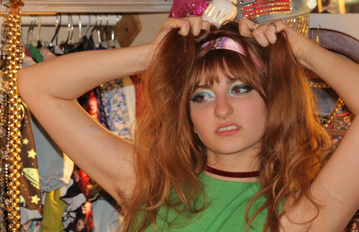 Isabella pulling at her hair in a green shift dress among racks of vintage clothes